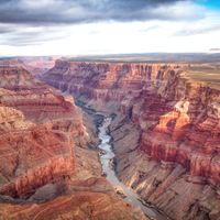 A wide view of the Colorado River winding through the layered red rock walls of the Grand Canyon under a cloudy sky.
